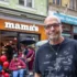 Starší muž bez vlasů a v tričku stojí venku na ulici a za ním jsou další lidé a podnik s bílým názvem nad vchodem