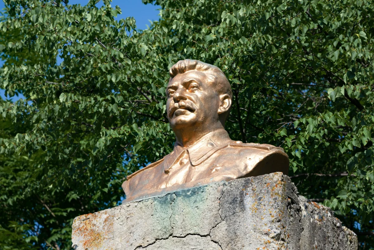 Bronze bust statue of a man atop a weathered stone pedestal, with green leafy trees and blue sky in the background.