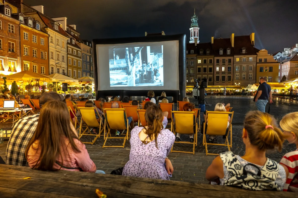 People seated in orange chairs watching a film on a large inflatable screen in a nighttime outdoor square with historic buildings surrounding them.