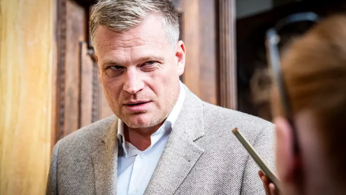 Muž ve středních letech ve světlém saku a světlé košili se dívá na někoho po svém levém boku