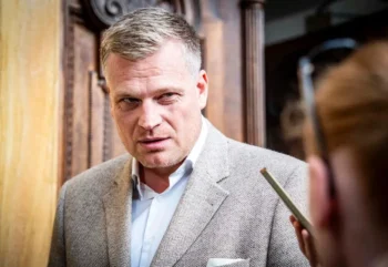 Muž ve středních letech ve světlém saku a světlé košili se dívá na někoho po svém levém boku