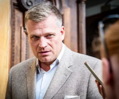 Muž ve středních letech ve světlém saku a světlé košili se dívá na někoho po svém levém boku