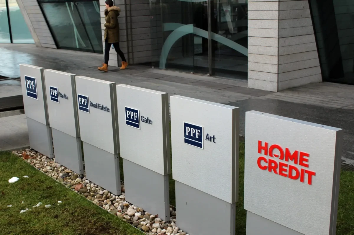 Row of grey sign panels with PPF logos and labels (Banka, Real Estate, Gate, Art) set outside a modern glass building; a person in a brown coat and orange boots walks by nearby.