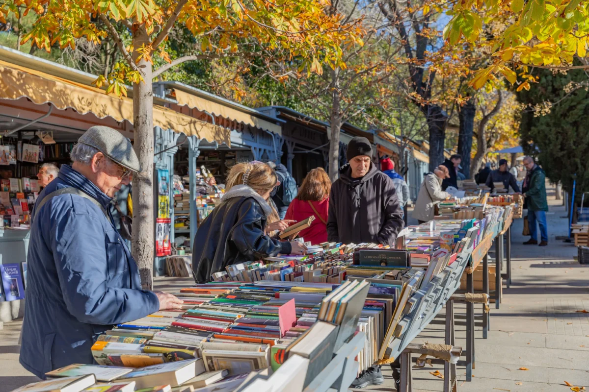 shutterstock_2689941013 1 | TVGURU.cz Outdoor street book market with tables full of books and shoppers browsing under autumn-leaved trees in sunlight.