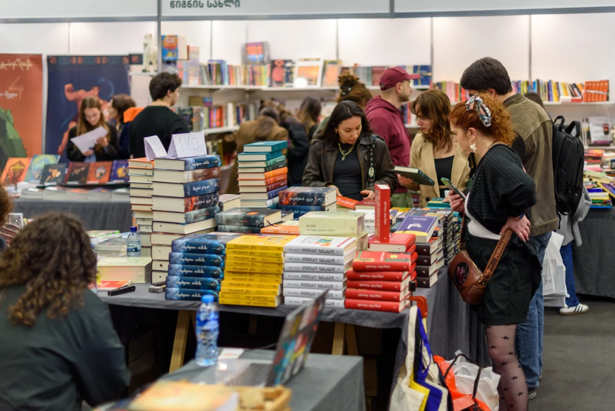 shutterstock_2771010941 1 | TVGURU.cz Crowded bookstore stall with stacks of colorful books on tables and shoppers browsing in the background.