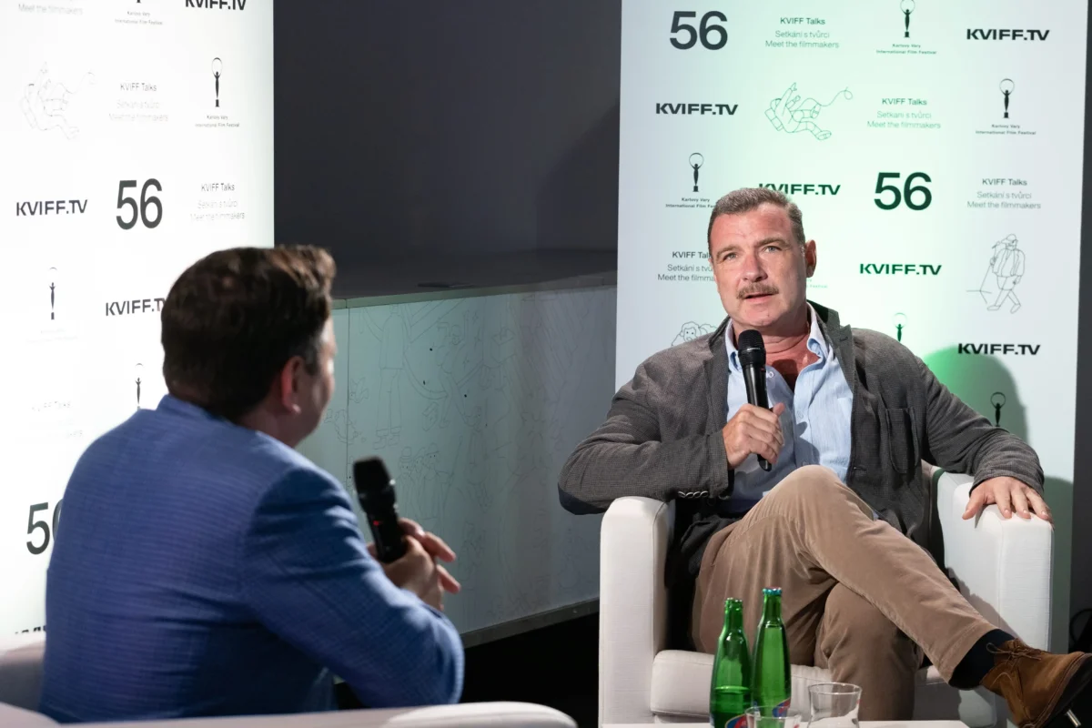 shutterstock_2175492811 1 | TVGURU.cz Two men in a panel interview on a stage, one speaking into a handheld microphone, with a branded backdrop and water bottles on the table.