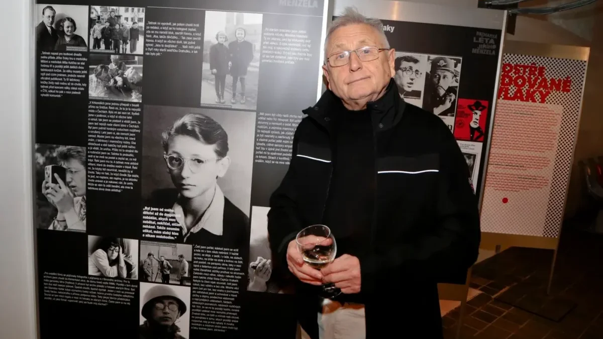 Older man with glasses in a black jacket stands next to a large exhibition panel about Jiri Menzel, holding a glass of wine.