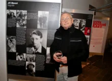 Older man with glasses in a black jacket stands next to a large exhibition panel about Jiri Menzel, holding a glass of wine.