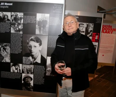 Older man with glasses in a black jacket stands next to a large exhibition panel about Jiri Menzel, holding a glass of wine.