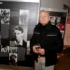 Older man with glasses in a black jacket stands next to a large exhibition panel about Jiri Menzel, holding a glass of wine.
