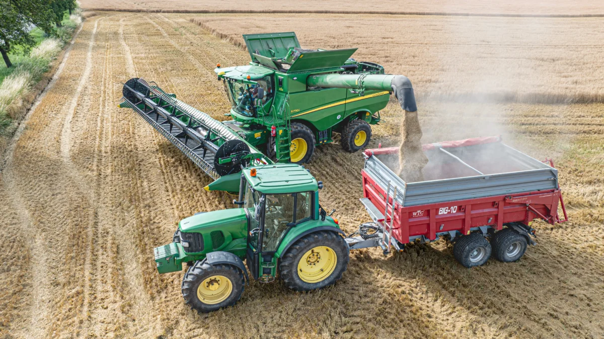 shutterstock_2487085523 1 | TVGURU.cz Green John Deere combine harvester unloading grain into a red trailer in a harvested field