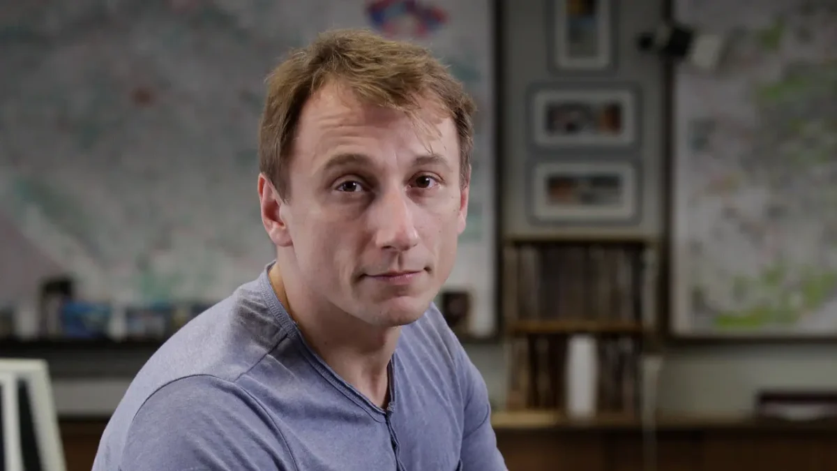 Close-up of a man in a light blue-gray T-shirt looking at the camera, with maps and framed photos on a wall behind him in a classroom or office setting.