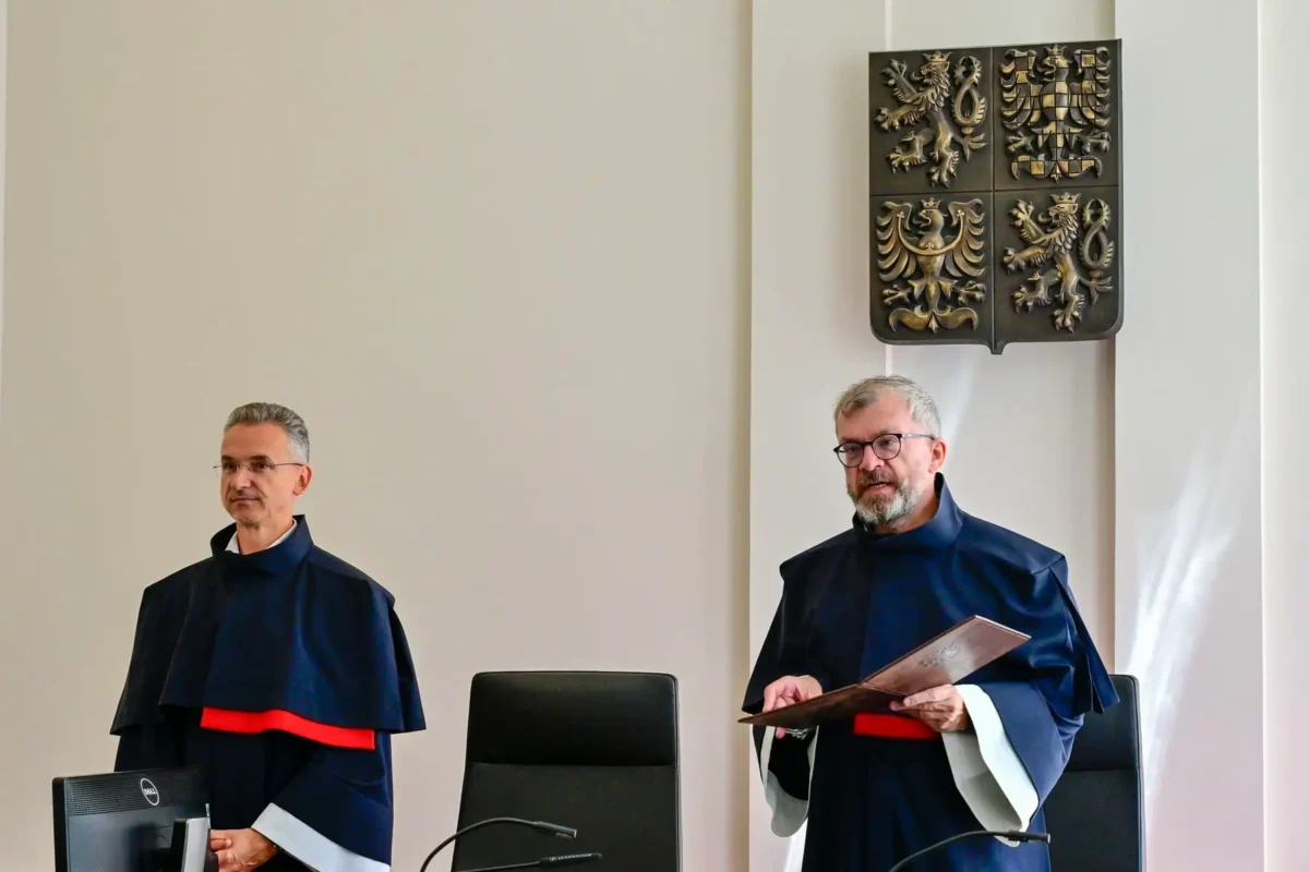 profimedia-1122477598 1 | TVGURU.cz Two judges in dark robes with red trim stand beside chairs in a courtroom; a four-panel heraldic shield hangs on the wall behind them.