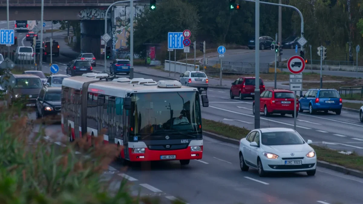 Red and white city bus dominates the foreground as cars line a busy urban intersection with traffic lights and signs nearby, under an overpass with graffiti.