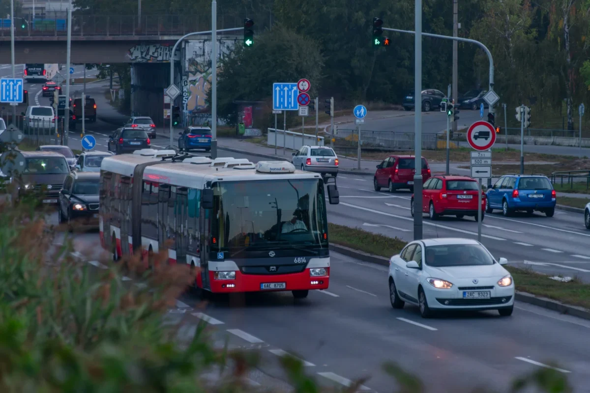 Red and white city bus dominates the foreground as cars line a busy urban intersection with traffic lights and signs nearby, under an overpass with graffiti.