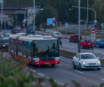Red and white city bus dominates the foreground as cars line a busy urban intersection with traffic lights and signs nearby, under an overpass with graffiti.