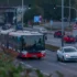 Red and white city bus dominates the foreground as cars line a busy urban intersection with traffic lights and signs nearby, under an overpass with graffiti.