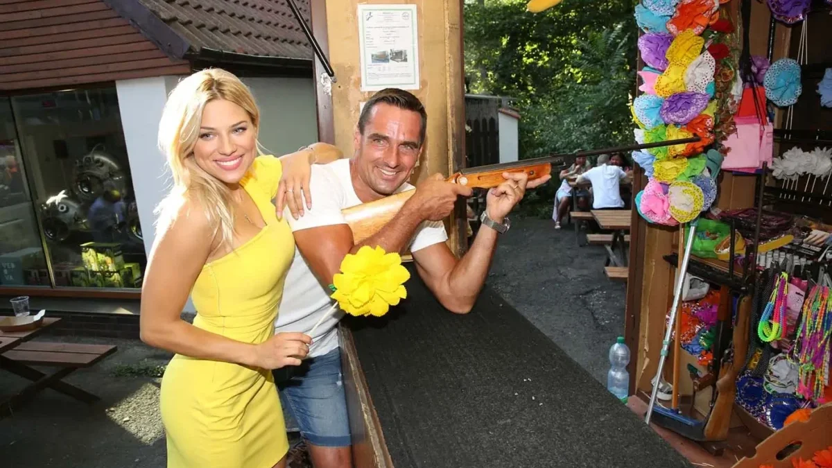 Žena s blond vlasy a ve žlutých šatech se směje a stojí vedle muže s krátkými vlasy, který v ruce drží zbraň