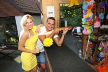 Žena s blond vlasy a ve žlutých šatech se směje a stojí vedle muže s krátkými vlasy, který v ruce drží zbraň