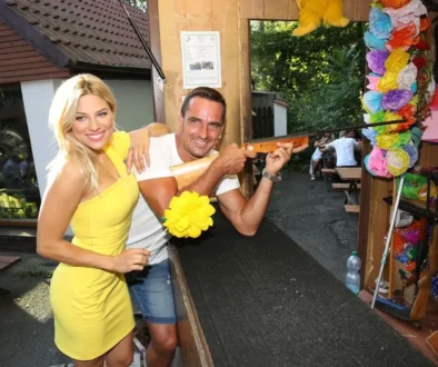 Žena s blond vlasy a ve žlutých šatech se směje a stojí vedle muže s krátkými vlasy, který v ruce drží zbraň