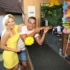 Žena s blond vlasy a ve žlutých šatech se směje a stojí vedle muže s krátkými vlasy, který v ruce drží zbraň