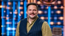Smiling man in a tan shirt and dark vest standing in a bright TV studio with blue lights behind him.