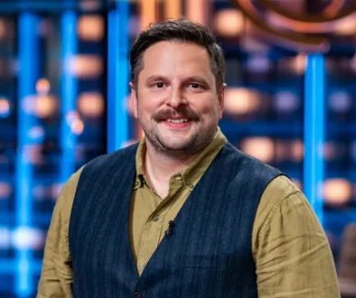 Smiling man in a tan shirt and dark vest standing in a bright TV studio with blue lights behind him.