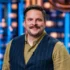 Smiling man in a tan shirt and dark vest standing in a bright TV studio with blue lights behind him.