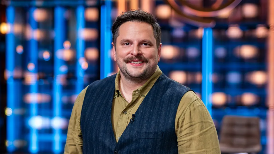 Smiling man in a tan shirt and dark vest standing in a bright TV studio with blue lights behind him.