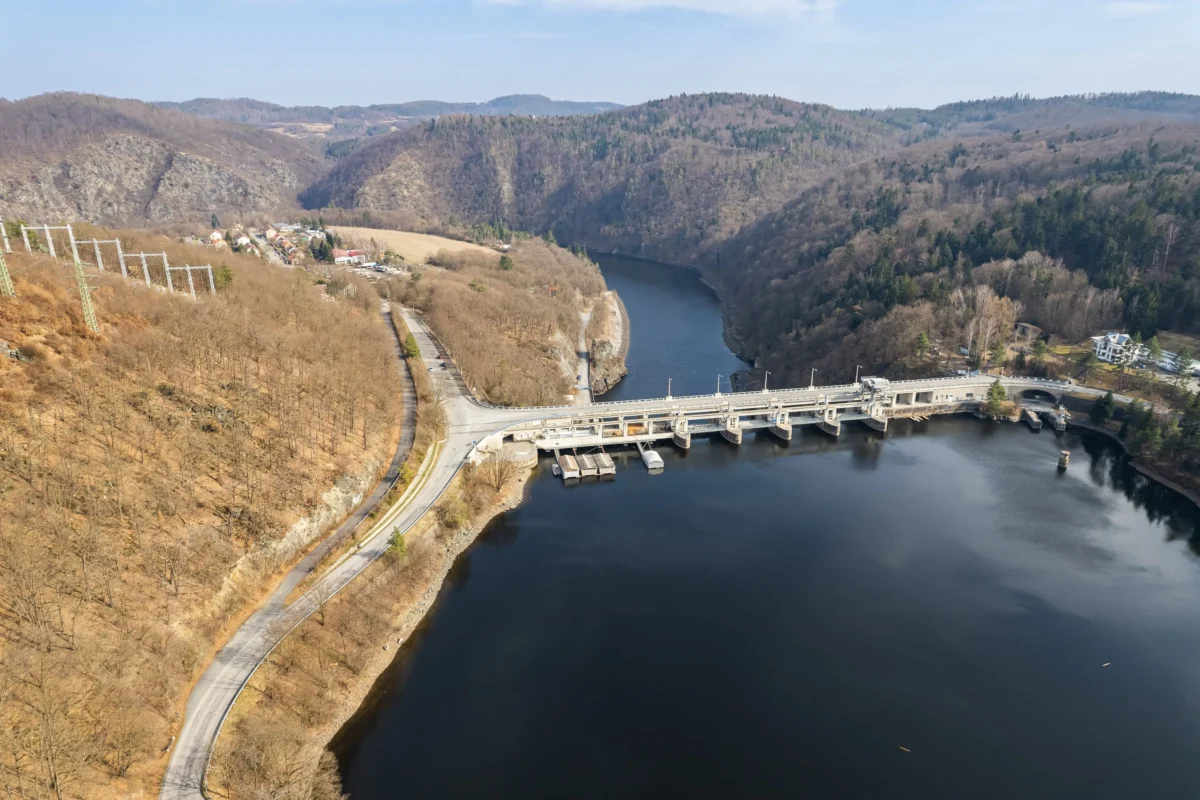 shutterstock_2568081603 1 | TVGURU.cz Aerial view of a dam and bridge spanning a wide river through a leafless, hilly valley with a small village nearby.