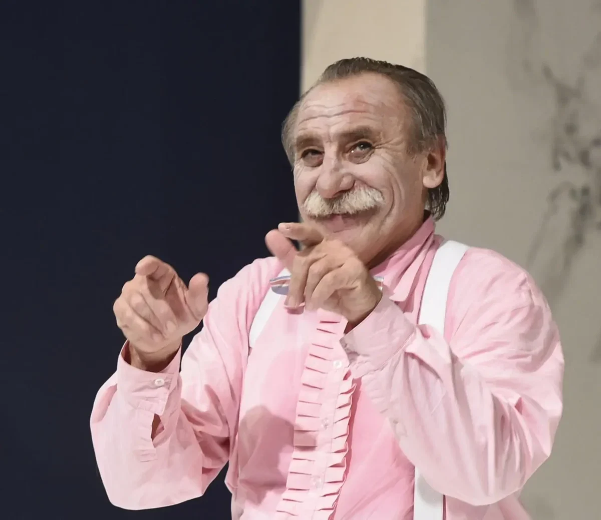 Older man with gray mustache in a pink shirt and white suspenders, smiling and clapping his hands toward the camera.