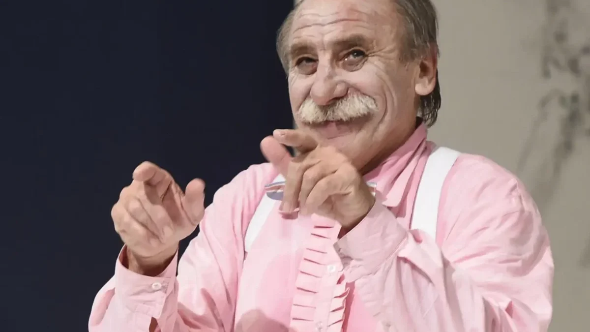 Older man with gray mustache in a pink shirt and white suspenders, smiling and clapping his hands toward the camera.
