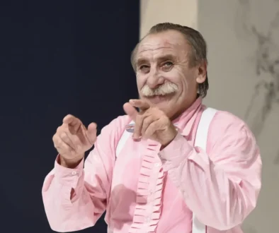 Older man with gray mustache in a pink shirt and white suspenders, smiling and clapping his hands toward the camera.