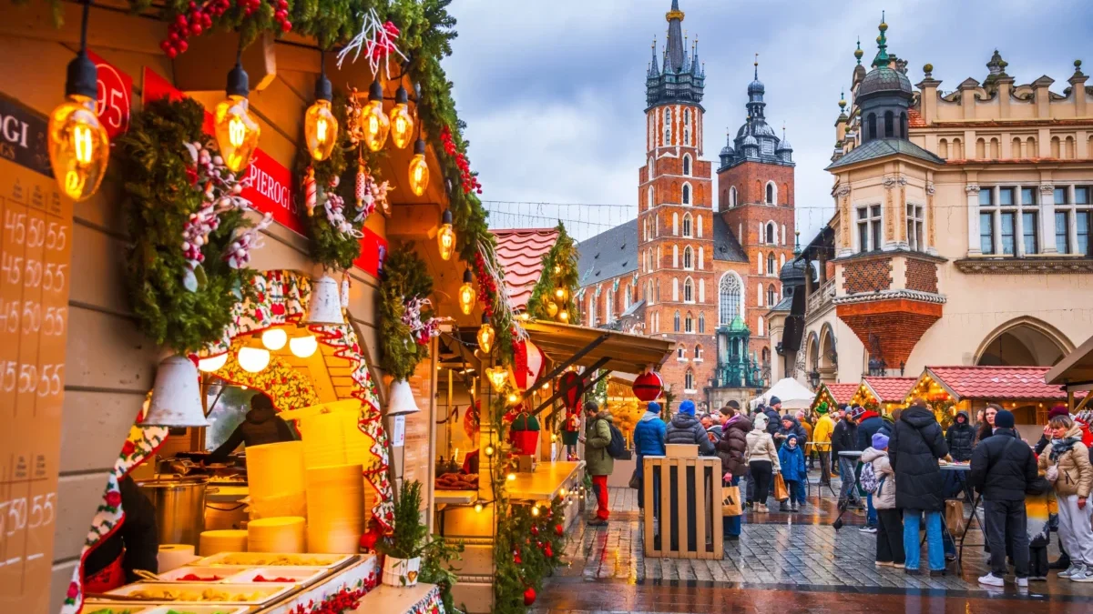 Bustling Christmas market with lit stalls, garlands, and shoppers on a wet cobblestone street; historic church towers rise in the background.