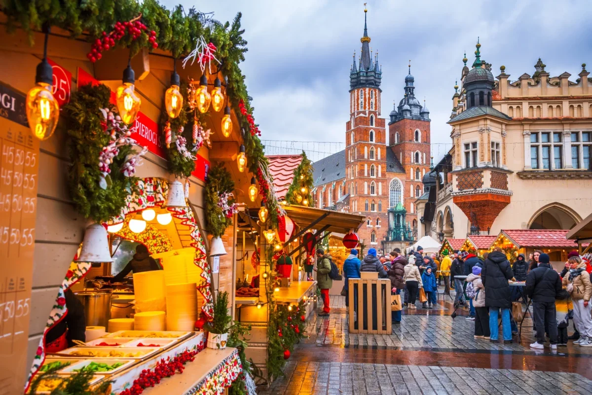 Bustling Christmas market with lit stalls, garlands, and shoppers on a wet cobblestone street; historic church towers rise in the background.