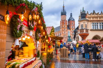 Bustling Christmas market with lit stalls, garlands, and shoppers on a wet cobblestone street; historic church towers rise in the background.