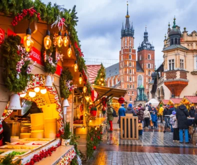 Bustling Christmas market with lit stalls, garlands, and shoppers on a wet cobblestone street; historic church towers rise in the background.