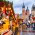 Bustling Christmas market with lit stalls, garlands, and shoppers on a wet cobblestone street; historic church towers rise in the background.