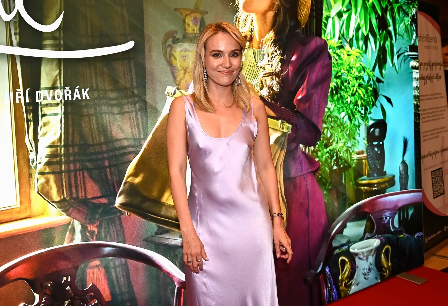 Woman in a lavender satin dress smiles at a colorful event backdrop behind her.