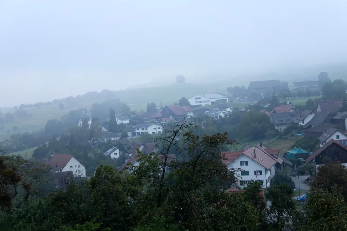 Foggy hillside village with red-roofed houses and trees fading into pale misty sky to the left and across the landscape.