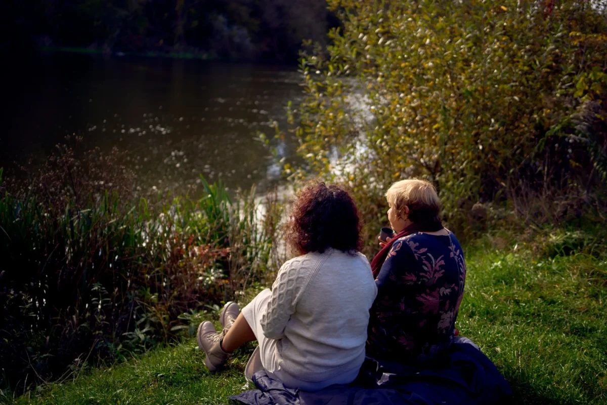 Two people sit close together on a blanket by a lake, seen from behind among tall grasses and autumn foliage.
