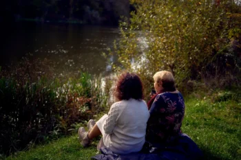Two people sit close together on a blanket by a lake, seen from behind among tall grasses and autumn foliage.