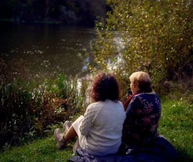 Two people sit close together on a blanket by a lake, seen from behind among tall grasses and autumn foliage.