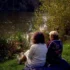 Two people sit close together on a blanket by a lake, seen from behind among tall grasses and autumn foliage.