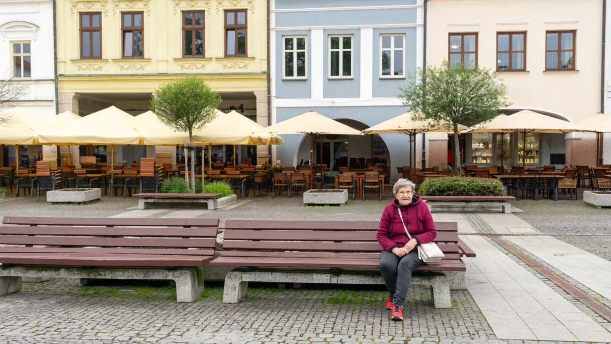 Elderly woman in a pink jacket sits on a bench in a cobblestone square with a cafe terrace and colorful facades behind her, beige umbrellas overhead.