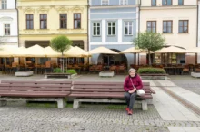 Elderly woman in a pink jacket sits on a bench in a cobblestone square with a cafe terrace and colorful facades behind her, beige umbrellas overhead.