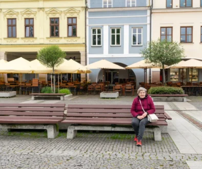Elderly woman in a pink jacket sits on a bench in a cobblestone square with a cafe terrace and colorful facades behind her, beige umbrellas overhead.