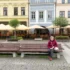 Elderly woman in a pink jacket sits on a bench in a cobblestone square with a cafe terrace and colorful facades behind her, beige umbrellas overhead.