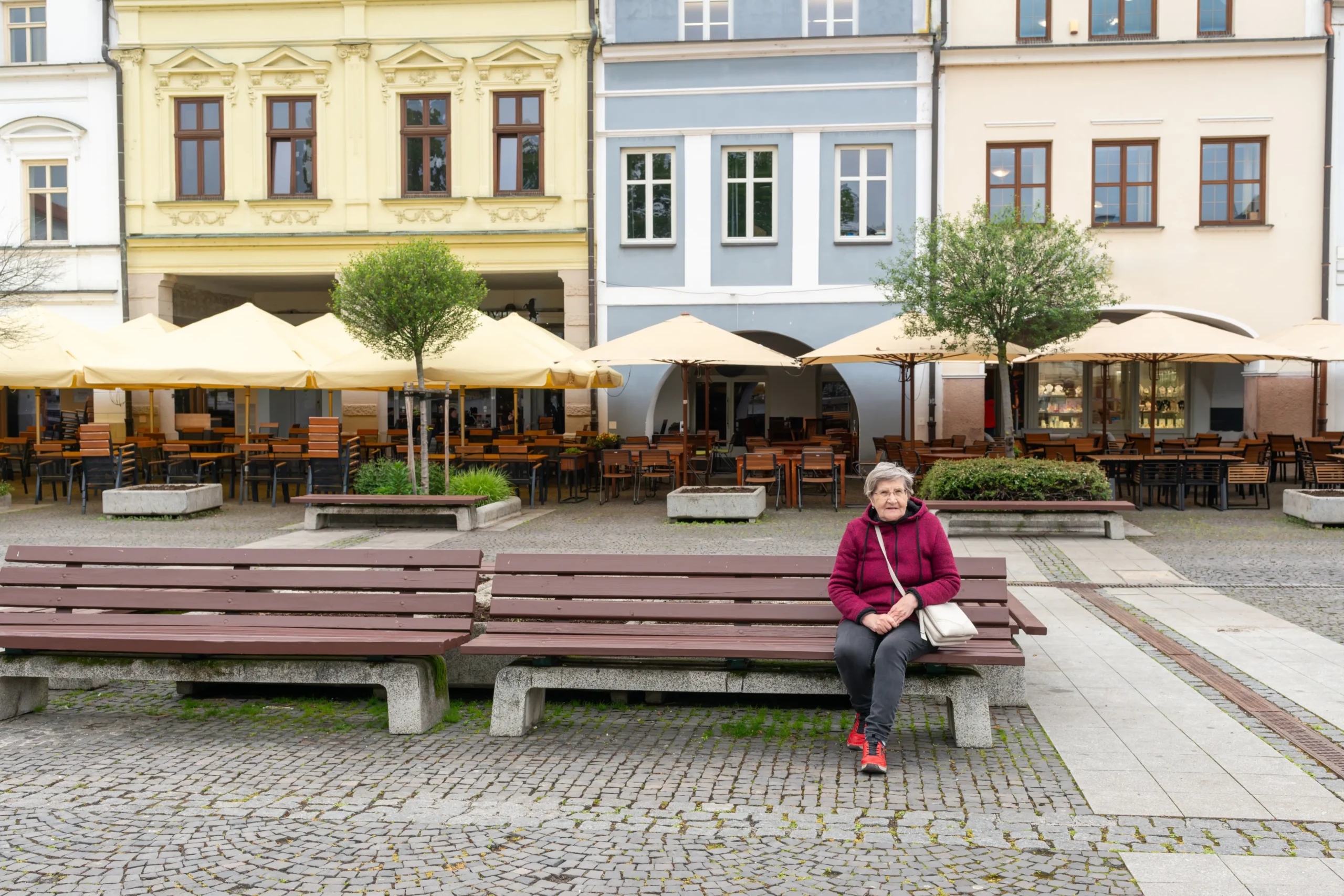Elderly woman in a pink jacket sits on a bench in a cobblestone square with a cafe terrace and colorful facades behind her, beige umbrellas overhead.