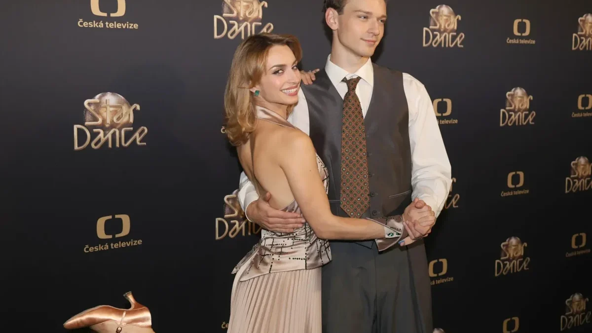 Smiling couple posing and dancing on a black backdrop with Star Dance and Česká televize logos; woman in a champagne dress and man in a gray vest and tie present on a red carpet backdrop.